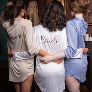 Puede incluir: Tres mujeres con pijamas de satén a juego con las palabras "Bride" y "Bridesmaid" impresas en la espalda. Las mujeres están de pie frente a una pared de madera oscura con un espejo enmarcado en oro y una lámpara de araña.