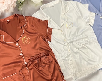 May include: Three satin pajama sets in different colors: rust, white, and light blue. Each set has a short-sleeved top with a button-up collar and matching shorts. The tops have a gold embroidered monogram with a floral design.