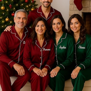 May include: A family wearing matching pajamas in shades of burgundy and green, posing in front of a Christmas tree and fireplace. The burgundy pajamas have white piping, and some are embroidered with names. The green pajamas also have white piping.