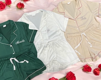 May include: Three sets of personalized pajama sets in green, white, and beige. Each set includes a short-sleeved button-up top and matching shorts. The tops are embroidered with names in cursive script. Red roses are scattered around the pajamas.