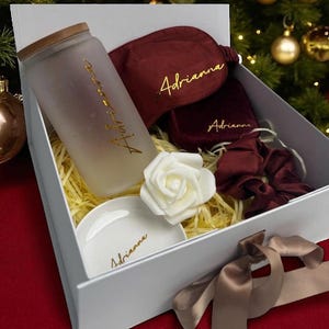 May include: A gift box containing a frosted glass tumbler with a wooden lid, a burgundy sleep mask, a small pouch, a scrunchie, a white rose, and a small white dish. The items are personalized with the name "Adrianna" in gold script.