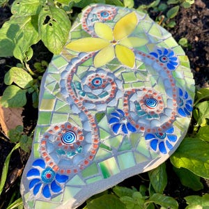 Könnte beinhalten: Ein Mosaik-Gartenstein mit einer gelben Blume und blauen floralen Akzenten. Der Stein besteht aus kleinen, farbigen Fliesen in Grün-, Blau- und Orangetönen. Der Stein ist von grünem Laub umgeben.