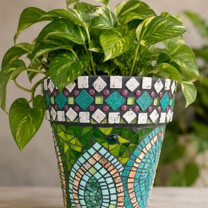 Può includere: Un vaso per piante conico decorato con un mosaico di tessere in vetro e pietra nei toni del verde, verde acqua e bianco. Il bordo superiore presenta un motivo geometrico di quadrati bianchi, rombi verde acqua e piccoli cerchi viola sul corpo.