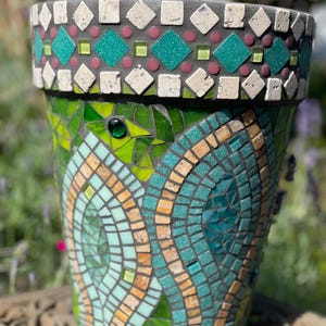 Op de afbeelding: Handgemaakte mozaïek bloempot met een decoratieve rand. De rand heeft witte ruitvormige tegels, turquoise vierkanten en roze cirkels. De pot is versierd met turquoise, groene en bruine mozaïektegels, die een ingewikkeld patroon vormen.