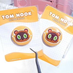 May include: Two Tom Nook button badges on orange backing cards. Each badge features a cartoon raccoon with dollar sign eyes. The cards read "TOM NOOK BUTTON BADGE" in white text. The website starwoodbay.com is printed at the bottom of the card.
