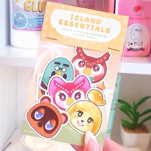 Puede incluir: Un paquete de pegatinas con personajes de Animal Crossing. El paquete incluye un oso marrón, un perro amarillo, un búho rosa, un pájaro verde y un gato rosa. Las pegatinas están en una bolsa de plástico transparente con un fondo amarillo y naranja. El texto "Island Essentials" está impreso en la bolsa.