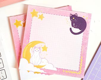 May include: Two pink and white gridded memo pads with a moon and cat design. The top pad features a black cat with a star on its forehead. The bottom pad features a white cat sitting on a crescent moon.