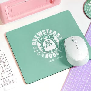 May include: A green mousepad with a white logo that says "Brewsters Roost - Est. 2005" with a cartoon bird wearing a bow tie. A white wireless mouse is on the mousepad.