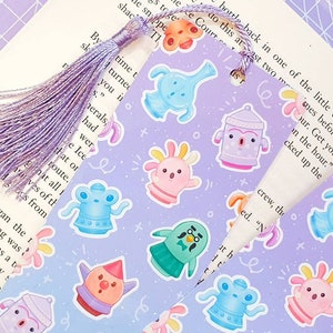 May include: A purple bookmark with a tassel and a pattern of colourful cartoon characters. The bookmark is folded over a page of a book with text that reads "Get your crew, the...mes before we...on the hour."