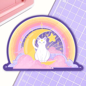 Puede incluir: Un gato blanco con una estrella en la frente está sentado en una luna creciente, rodeado de un arcoíris y nubes. El fondo es de color morado oscuro con estrellas y brillos.