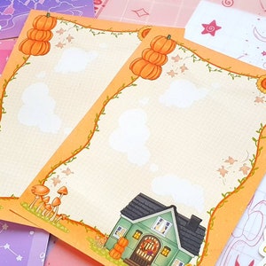 May include: Two blank orange and white printable note cards with an autumn theme. The cards feature a vine border with pumpkins, mushrooms, and a small house with a wreath on the door.