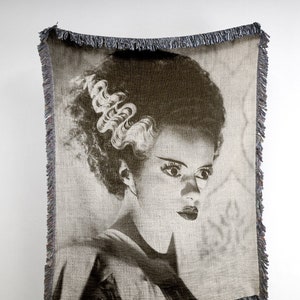 Puede incluir: Una manta tejida en blanco y negro con un retrato de la Novia de Frankenstein. La imagen es un primer plano de su rostro y su cabello.