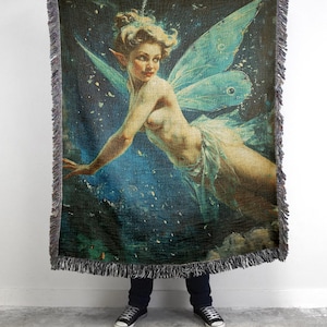 May include: Woven throw blanket featuring a vintage illustration of a fairy with blue wings against a dark blue and starry background. The blanket has a fringed edge and is held up by a person wearing black shoes and jeans.