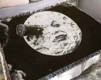 May include: A black and white woven throw blanket with a moon face looking through a telescope. The moon face is smiling and has a large nose.