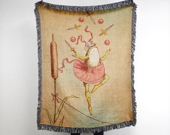 May include: A woven tapestry featuring a green frog wearing a pink tutu and juggling red balls and silver knives. The frog is against a beige background with a brown reed in the corner.