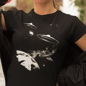 May include: A black t-shirt with a white graphic of a planet and three flying saucers in space. The graphic is stylized and has a vintage feel.