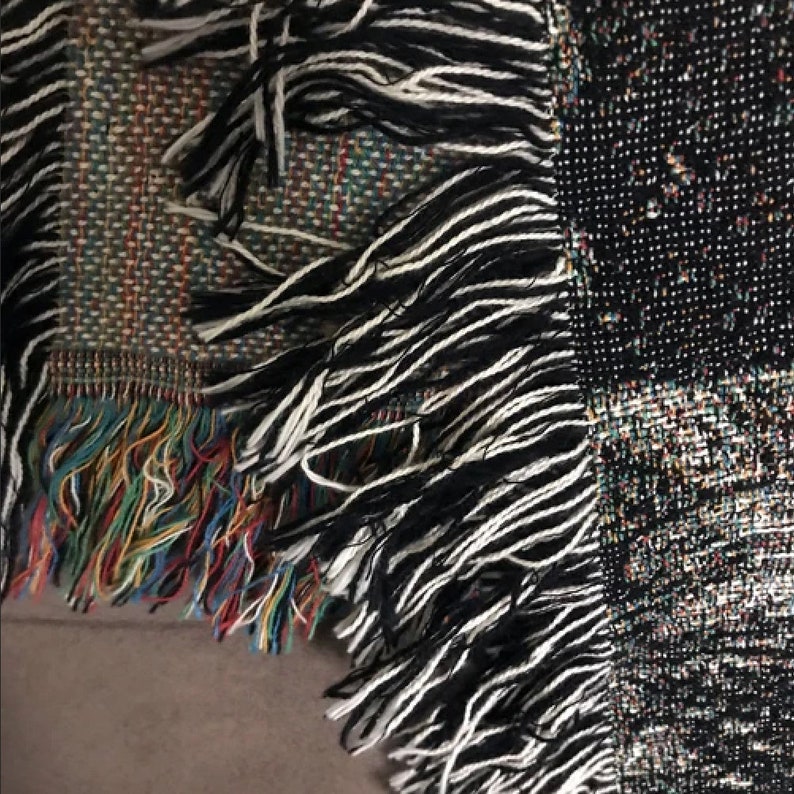 Op de afbeelding: Close-up van een geweven deken met zwarte, witte en veelkleurige franjes. De deken heeft een patchwork-ontwerp met verschillende patronen en kleuren.
