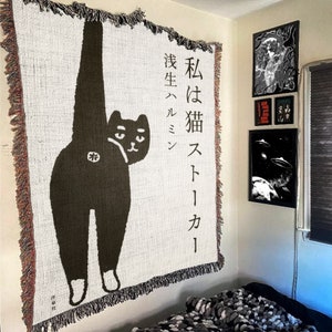 May include: A white tapestry with a black cat illustration and Japanese text that reads "I am a cat, born to be cool". The tapestry has a fringe border.