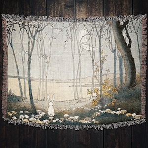 May include: A woven tapestry featuring a white rabbit standing in a forest with a moon in the background. The forest is depicted with tall, thin trees and a river in the distance. The tapestry has a fringed border.