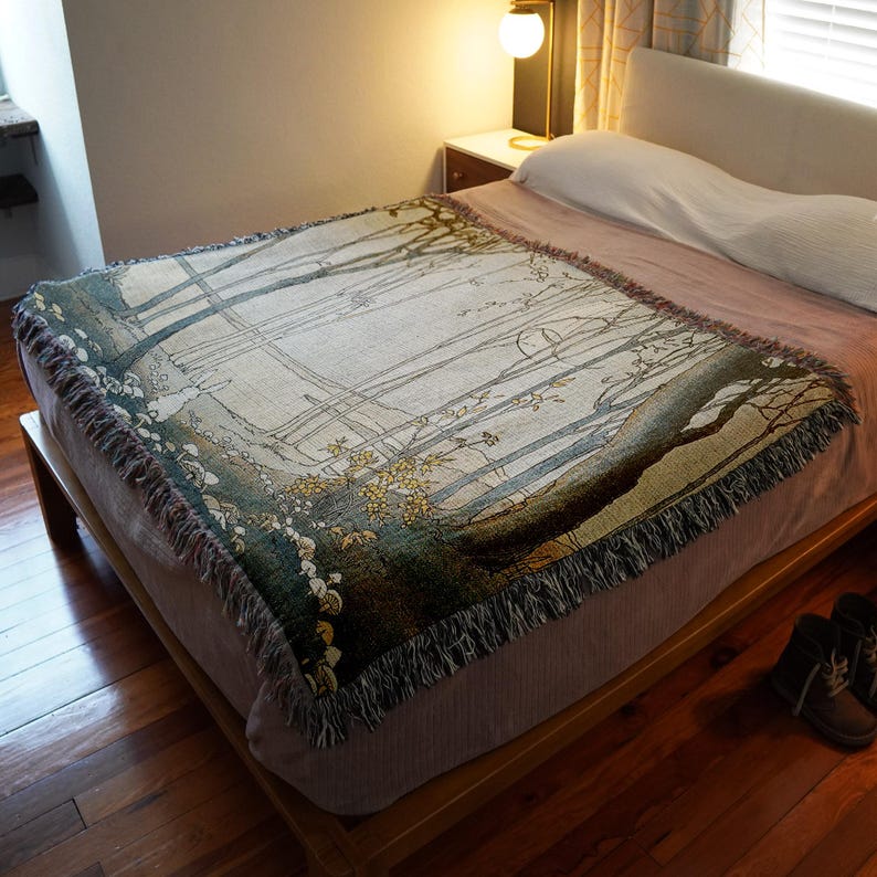 May include: A woven tapestry throw blanket with a forest scene featuring trees, branches, and leaves in shades of green, brown, and white. The blanket has a fringed edge and is draped over a bed.