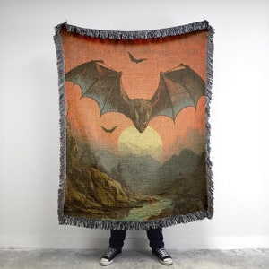 May include: A woven tapestry throw blanket with a vintage illustration of a bat flying in front of a sunset. The bat has large wings and is flying over a landscape with a river and mountains.