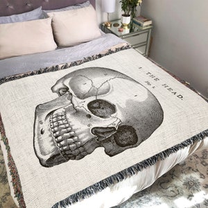 May include: A woven throw blanket with a black and white anatomical illustration of a human skull. The skull is labeled with numbers and the text "THE HEAD."