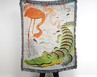 May include: A woven tapestry featuring a green alligator with red spots, sleeping on a white background with pink and orange flamingos.