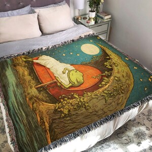 May include: A woven tapestry throw blanket with a whimsical design featuring a green frog sleeping on a crescent moon. The background is a dark blue with stars and a full moon.