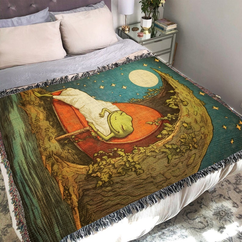 May include: A woven tapestry throw blanket with a whimsical design featuring a green frog sleeping on a crescent moon. The background is a dark blue with stars and a full moon.