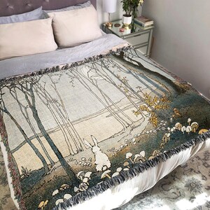May include: A woven tapestry throw blanket with a forest scene featuring a white rabbit and mushrooms. The blanket has a light blue background with brown trees and a light brown river. The blanket has a fringe border.