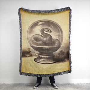 Könnte beinhalten: Eine gewebte Wanddecke aus Tapisserie mit einer Vintage-Illustration einer Schlange in einer Glaskugel auf einem Sockel. Der Hintergrund ist in einem gedämpften Gelb mit einem braunen und weißen Design gehalten.
