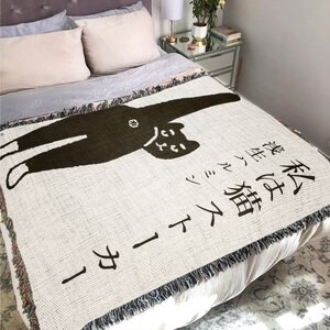 May include: A white throw blanket with a black cat illustration and Japanese text that reads "This cat is a little bit of a rascal".