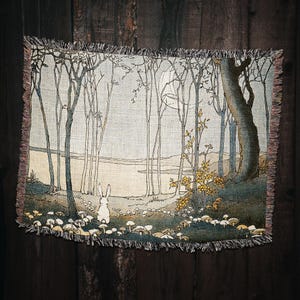 May include: A tapestry depicting a white rabbit standing in a forest with a moon in the sky. The forest is made up of tall, thin trees with a river in the background. The rabbit is surrounded by small, white mushrooms.