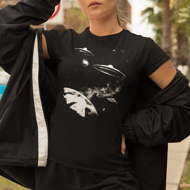 May include: A black t-shirt with a white graphic of a planet and three flying saucers in space. The graphic is stylized and has a vintage feel.