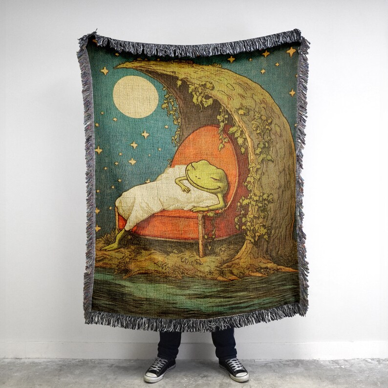 May include: A woven tapestry throw blanket featuring a green frog sleeping in a red armchair under a crescent moon and stars. The frog is wearing a white blanket. The background is a dark blue with a green tree and a river.
