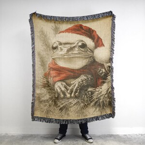 May include: A woven throw blanket featuring a cartoon illustration of a frog wearing a Santa hat and a red scarf. The frog is sitting in a pile of what appears to be snow.