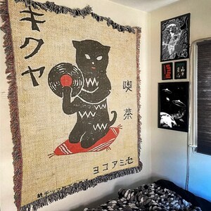 May include: A beige tapestry with a black cat holding a vinyl record. The cat is sitting on a red cushion. The tapestry has Japanese text that reads "キワヤ" and "喫茶". The bottom of the tapestry reads "ヨコハマモモ".