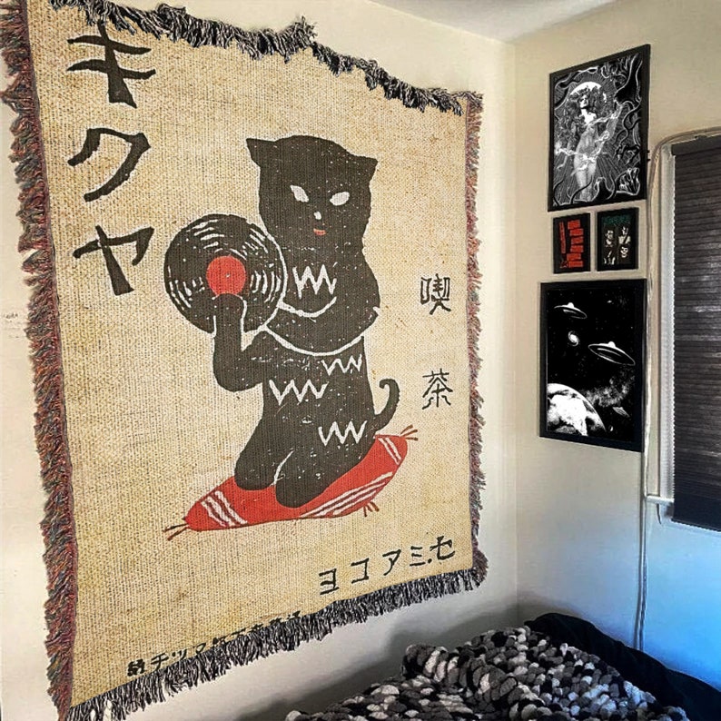 May include: A beige tapestry with a black cat holding a vinyl record. The cat is sitting on a red cushion. The tapestry has Japanese text that reads "キワヤ" and "喫茶". The bottom of the tapestry reads "ヨコハマモモ".