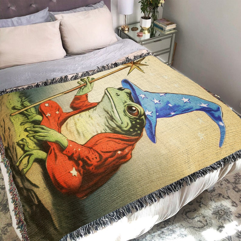 Op de afbeelding: Een geweven wandkleed deken met een fantasievolle illustratie van een groene kikker die een blauw-witte tovenaarshoed draagt en een toverstaf vasthoudt. De kikker draagt een rood-oranje gewaad met gouden sterren. De achtergrond is lichtbeige.
