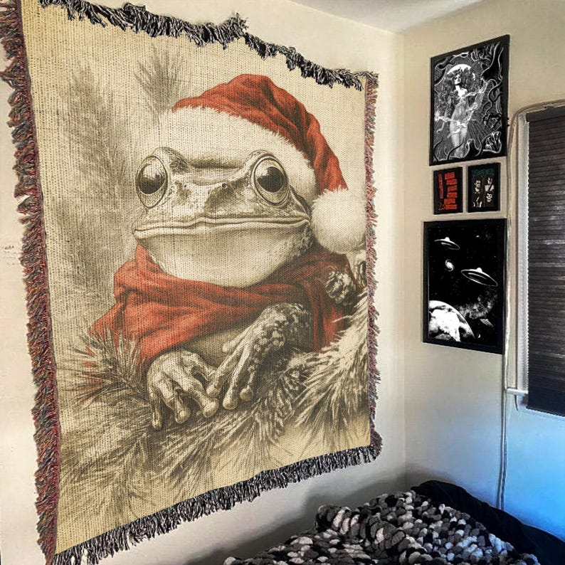 May include: A tapestry featuring a frog wearing a Santa hat and a red scarf. The frog is sitting in a pile of brown and white fur. The tapestry has a fringe border.