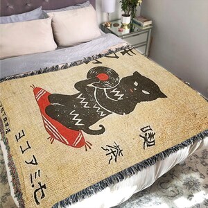 May include: A tan throw blanket with a black cat holding a red record. The cat is wearing a white and black patterned sweater. The blanket has Japanese text that reads "おにぎり" and "お茶".