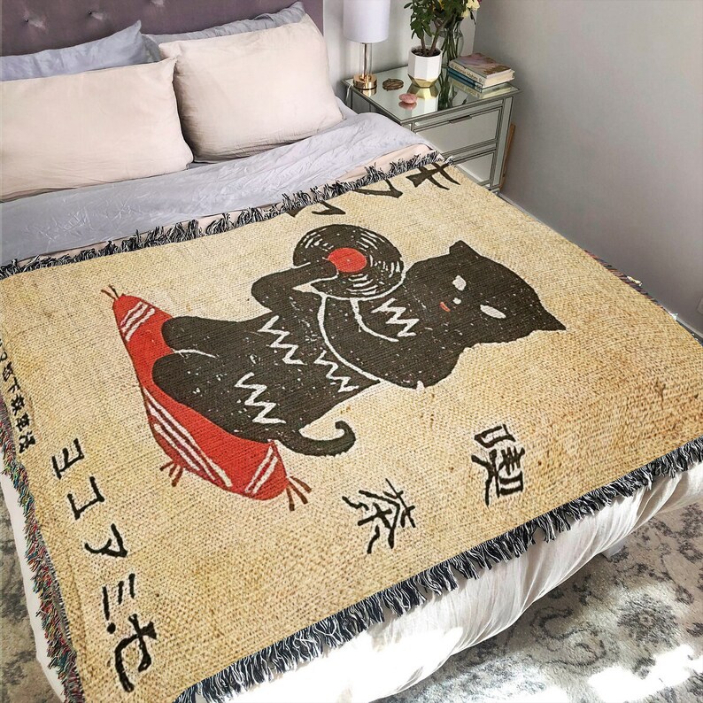 May include: A tan throw blanket with a black cat holding a red record. The cat is wearing a white and black patterned sweater. The blanket has Japanese text that reads "おにぎり" and "お茶".