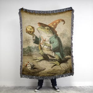 May include: Woven tapestry throw blanket featuring a frog dressed as a wizard, holding a twenty-sided die. The frog wears a green coat and a pointed hat. A six-sided die is on the ground. The blanket has a fringed edge.
