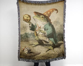 May include: Woven tapestry throw blanket featuring a frog dressed as a wizard, holding a twenty-sided die. The frog wears a green coat and a pointed hat. A six-sided die is on the ground. The blanket has a fringed edge.