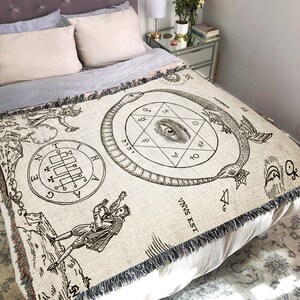 Puede incluir: Una manta de tejido de tapiz con fondo beige e ilustraciones en blanco y negro de una serpiente, un ojo y un pentagrama. El texto "VNVS EST" es visible en la esquina inferior derecha.