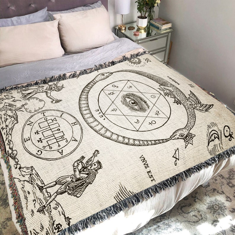 Puede incluir: Una manta de tejido de tapiz con fondo beige e ilustraciones en blanco y negro de una serpiente, un ojo y un pentagrama. El texto "VNVS EST" es visible en la esquina inferior derecha.