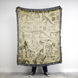 May include: A woven throw blanket featuring a vintage illustration of various dinosaur skeletons in black and white on a beige background.