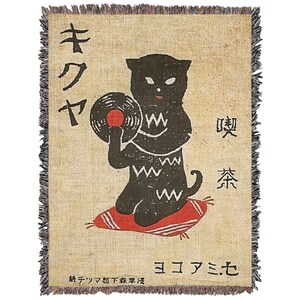 May include: A black cat holding a vinyl record with a red label, sitting on a red cushion. The cat is wearing a black and white patterned shirt. The background is a beige woven fabric with Japanese text that reads "キクヤ" and "喫茶".