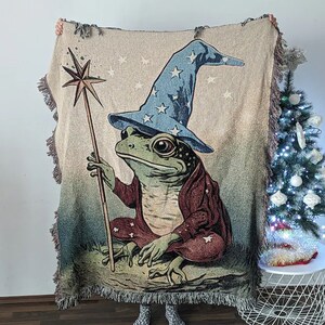 Op de afbeelding: Een geweven tapijtdeken met een groene kikker die een blauwe tovenaarshoed draagt en een toverstaf vasthoudt. De achtergrond is beige met sterren.
