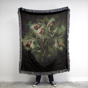 May include: A rectangular woven throw blanket featuring a dark black background with a central botanical illustration of bell-shaped flowers and dark berries in shades of green, brown, and purple, finished with a decorative fringed edge on all sides.